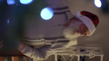 Dikey video, yakın plan, sakallı yaşlı bir adam Noel şapkası takıyor, dans ediyor.
