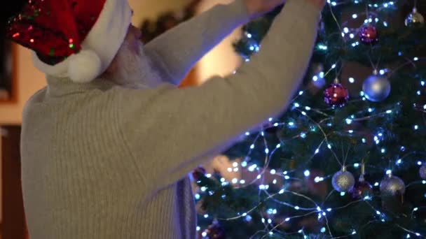 Пожилой Человек Красной Рождественской Шляпе Настраивает Гирлянду Елку — стоковое видео