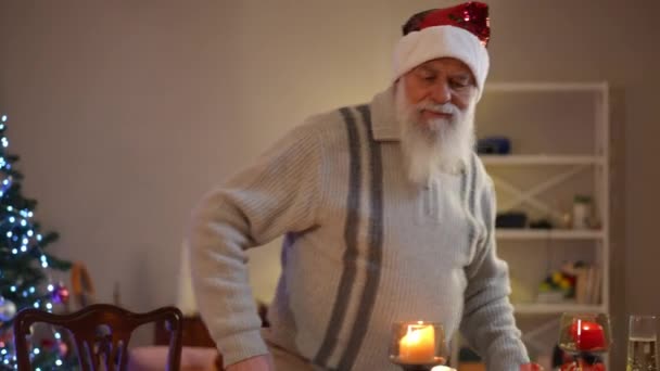 ひげでクリスマスの帽子をかぶった老人がお祝いのテーブルに座ります テーブルに座って周りを見回すと 男は彼の頭の上に手を置く — ストック動画