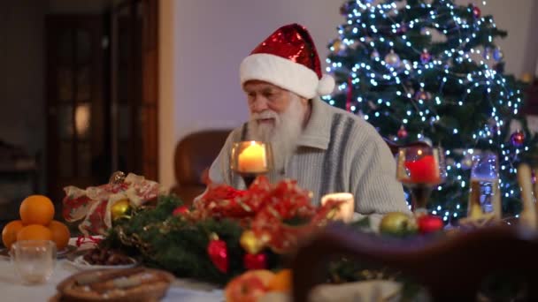 一位头戴圣诞礼帽的大胡子老人坐在圣诞树旁的喜庆桌旁 检查礼品盒 却没有发现礼品盒里有什么礼物 他从桌上站起来去找 — 图库视频影像