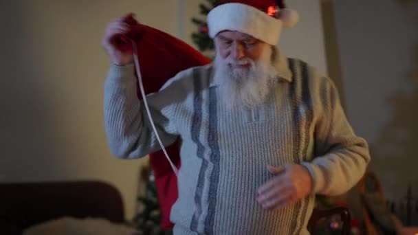 一个头戴圣诞礼帽的大胡子老人拿着一袋礼物 坐在桌旁 匆匆摘下礼帽 松了口气 — 图库视频影像