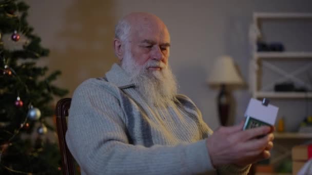 Пожилой Бородатый Мужчина Возле Елки Осматривает Игрушечный Домик Картона — стоковое видео