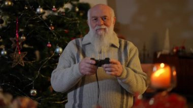 Noel ağacının yanında yaşlı sakallı bir adam iki eliyle kumanda kolunu kontrol ediyor ve ekrana bakıyor.