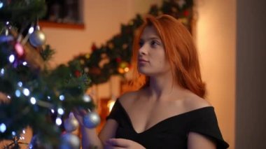 Noel ağacına bir Noel ağacı oyuncağı astıktan sonra genç kızıl saçlı bir kadın doğrudan kameraya bakar.