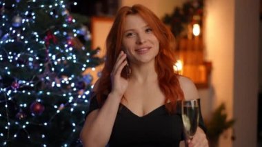Genç, güzel, kızıl saçlı kadın elinde bir bardak beyaz şarapla Noel ağacının yanında dururken cep telefonundan tebrik alıyor.