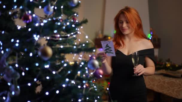 カクテルドレスの美しい若い女性は 彼女の手にシャンパンのグラスとクリスマスツリーの近くに立って 新しい年のカードを読みます シャンパンを飲むことは願い事の伝統です — ストック動画