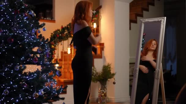 クリスマスツリーの近くに立って 美しい若い女性は大きな床の鏡で自分自身を見ています 鏡を見て 女性は彼女のカクテルドレスと笑顔を調整する — ストック動画