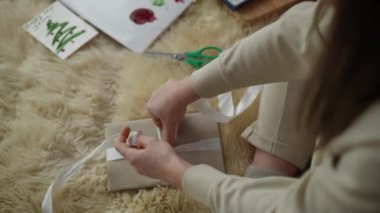 Bir kadın yerde otururken beyaz bir kurdeleyle hediye bağlıyor. Noel ve Yeni Yıl için hediye paketi