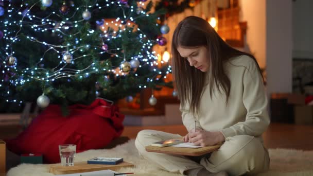 クリスマスツリーの下に座っている若い女性は グラファイト鉛筆で未来の新年のカードのスケッチを作ります クリスマスと新年の前夜のホームレジャー — ストック動画