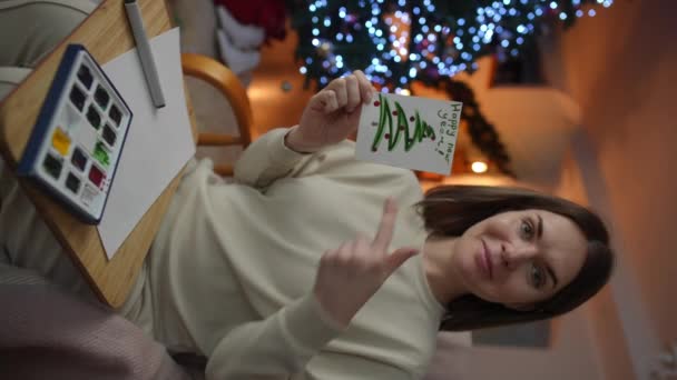 バーティカルビデオ ロッキングチェアに座っている女性は 手作りのカードを手に持って オペレータに新年のお祝いに参加するように誘います — ストック動画