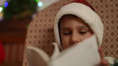 Noel şapkası takan bir çocuk kitap okur ve Noel 'de sallanan sandalyede otururken şiir öğrenmeye çalışır.