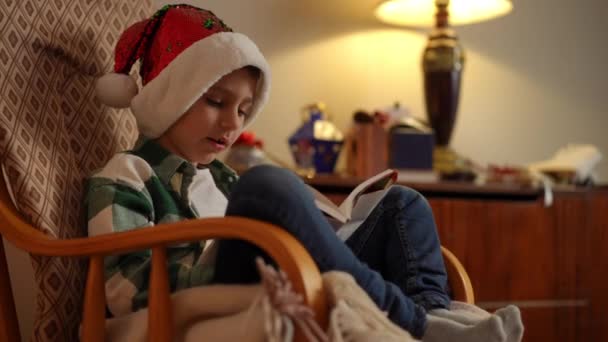 一个戴着圣诞礼帽坐在摇椅上的男孩笑着翻阅着一本书 — 图库视频影像