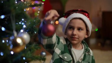 Noel ağacının yanında duran Noel şapkalı bir çocuk dalına güzel bir Noel topu asmaya çalışıyor.
