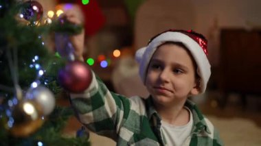Noel ağacı süslemekten doğan bir çocuğun yüzündeki mutluluk. Noel şapkalı bir çocuk Noel topunu dallara astığı için gülümsüyor.