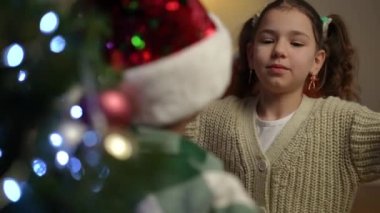 Noel ağacının yanında duran bir kız, Noel şapkalı bir çocuğa sarılıyor. Oğlanı kucaklayan kız, kameraya bakıp gülümsüyor.