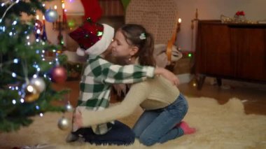 Noel ağacının yanında halının üzerinde karşılıklı otururken, kız ve erkek kardeşler birbirlerine sevinçle sarılıyorlar.