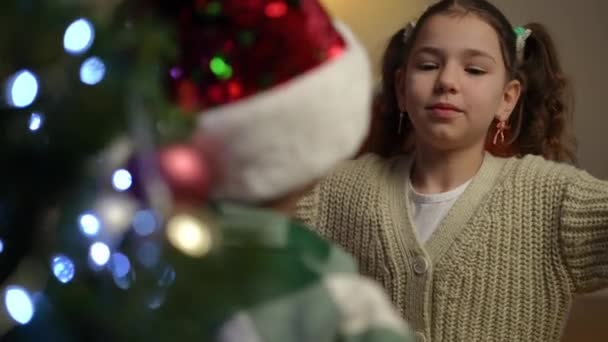 一个女孩站在圣诞树旁抱着一个戴着圣诞礼帽的男孩 女孩拥抱着男孩 对着镜头笑了 — 图库视频影像