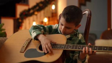 Bir çocuk elinde gitarla sandalyeye oturur ve onu çalmaya çalışır.