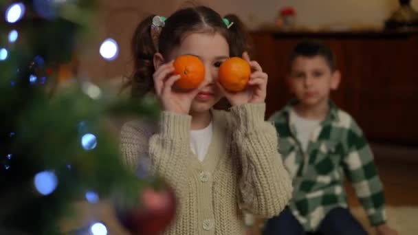 一个女孩站在圣诞树旁 一边看着摄像机一边玩 一边玩着两个橘子 — 图库视频影像