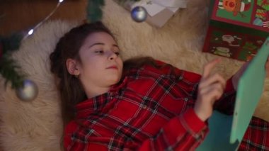 Noel sabahı uyumu ağır çekimde. Bir kız Noel ağacının yanında bir tabletle yatıyor ve bir Noel topuyla oynuyor.