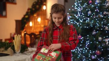 Noel için dekore edilmiş bir evde, Noel ağacının yanında duran bir kız elinde Noel hediyesi ile büyük bir kutu çevirir.
