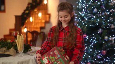 Elinde Noel hediyesi olan büyük bir kutuya bakarken, Noel ağacının yanında duran bir kız kutunun içindekileri tahmin etmeye çalışır.