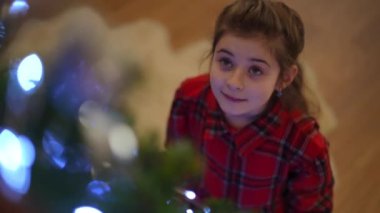 Evdeki bir kız Noel ağacına hayran, onun yanında duruyor.