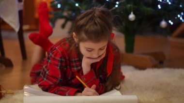Noel ağacının yanındaki evde Noel tatili. Kız halıya uzanıp resim çiziyor.