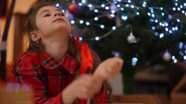 Oturma odasında bir kız Noel ağacının yanındaki halıya uzanmış, yüzünü ellerine yaslamış, kameraya bakıyor ve gülümsüyor.