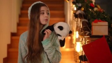 Pijamalı ve omuzunda oyuncak ayı olan bir kız yatağa gitmek istemez ama güzel dekore edilmiş bir merdivenin önünde Noel 'i beklemek ister.