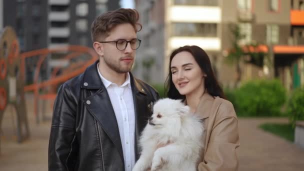 在现代住宅区的背景下 一对年轻漂亮的情侣抱着一只狗进行了特写 一个女人怀里抱着一只白色的狗 这对夫妇看着摄像机 — 图库视频影像