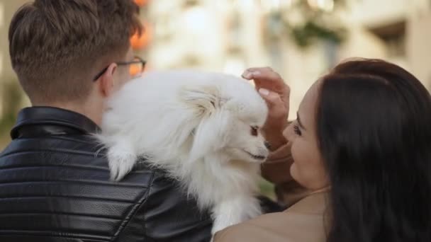 后视镜一对抱着狗的年轻夫妇 一个男人站在一个女人旁边 怀里抱着一只白狗 那只狗舔了一下女人的鼻子 直接看着摄像机 — 图库视频影像