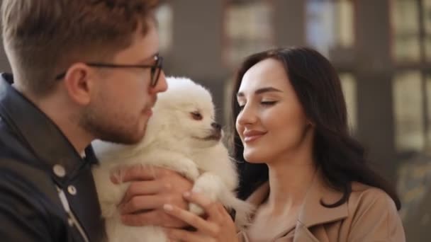 一对抱着狗的年轻夫妇 一个男人站在一个女人旁边时 怀里抱着一只白色的狗 一个男人和一个女人同时亲吻两边的一条白狗 — 图库视频影像