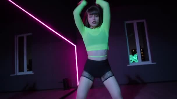 现场摄像头 一个身穿亮丽舞台服装的年轻漂亮的姑娘在一个有霓虹灯照明的黑暗的舞蹈大厅里跳舞 — 图库视频影像