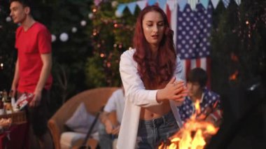 Kız ayakta duruyor, ellerini ateşin yanında ısıtıyor ve kameraya bakıyor. Açık havada arkadaşlarla barbekü.
