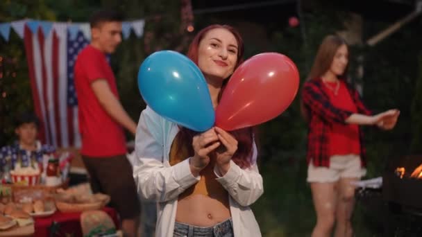 前景では 女の子が7月4日の独立記念日に友人の会社で風船を楽しんでいます アウトドアバーベキュー 少女は赤と青の風船を投げ カメラにキスを吹く — ストック動画