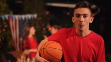 Yakın plan. Kolunun altında basketbol topu olan bir adam kameraya bakar ve arka plandaki bir partide sakız çiğner.