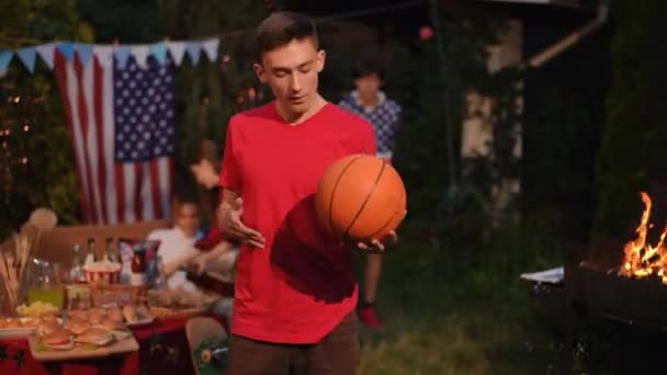 前景では 男は彼の手にバスケットボールで遊んで カメラを見ています 屋外バーベキューでの友人とのレジャータイム — ストック動画