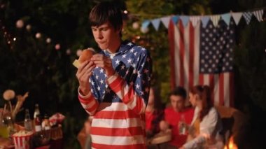 Bir genç arkadaşlarıyla açık hava partisinde seve seve hamburger yer. Bağımsızlık Günü için Amerikan yemekleri