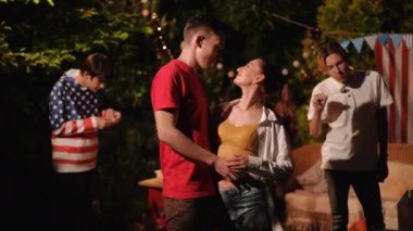 Bir adam bir kıza sarılıyor ve arkadaşlarıyla bir partide dikilirken gözlerinin içine bakıyor. Adam kameraya bakıyor ve arkadaşları çubuklarda marşmelov yiyor..