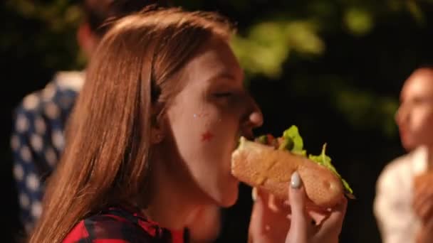 クローズアップ 少女はマスタードで寛大に汚染されたホットドッグを食べ 音楽のビートに踊ります 屋外パーティーで友達とストリートフード — ストック動画