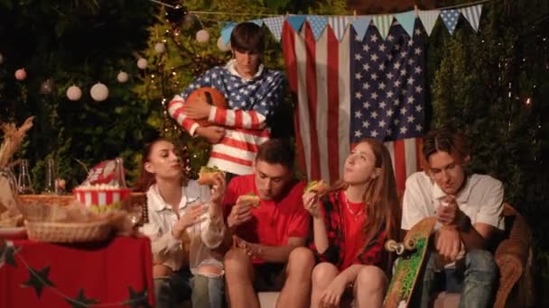 屋外のパーティーに座っている友人のグループは 自分で調理したホットドッグを食べます 7月4日のお祝いのコンセプト — ストック動画
