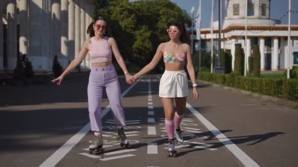 两个戴眼镜溜冰鞋的女朋友在一个城市公园里 在阳光灿烂的日子里 女孩子们在户外骑车牵着手 — 图库视频影像