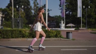 Gözlüklü güzel bir kadın güneşli bir günde şehir parkının arka planında patenle zarif bir şekilde hareket eder. Yavaş çekim