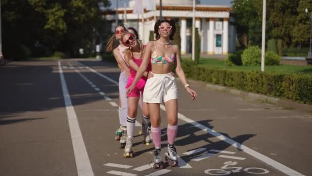 ローラーのスケートでサングラスで4人の若い女性がお互いを抱き合い 晴れた日に街の公園を通ってビートに移動します スローモーション — ストック動画