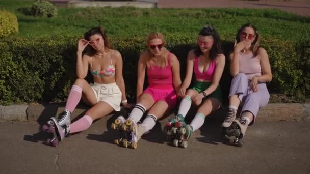 サングラスとローラースケートの4人の美しい若い女性は 明るく晴れた日に街の公園に座っています 女性は不思議に微笑み カメラを見る — ストック動画