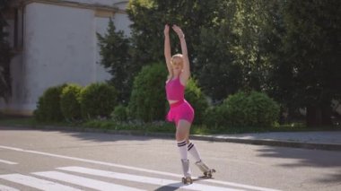 Güneş gözlüklü, patenli güzel bir kız güneşli bir günde şehir parkında dans figürleri sergiliyor. Kız tamamlanmış dans öğesine seviniyor..