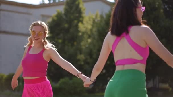 サングラスで踊る2人の少女が 街の公園で晴れた日に手をつないで踊る 自然の中で踊る女の子の顔の喜び — ストック動画