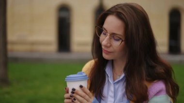 Sabahın erken saatlerinde, parkta bir bankta oturan bir kadın, bir bardaktan kahve içer ve bundan zevk alır. Yakın plan.