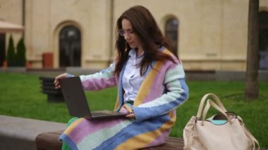 Gözlüklü genç bir kadın dizüstü bilgisayarını açar ve sabah şehir parkında bir bankta otururken çalışmaya, esnemeye başlar.